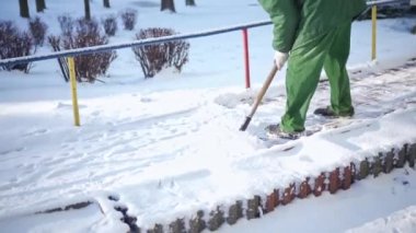A snowy alley of a pedestrian walkway in a city housing development. A man scrapes and tosses aside compacted snow. Morning work in winter.