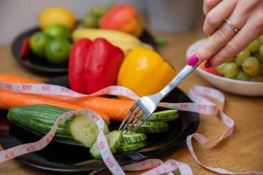 A plate with sliced cucumber and other vegetables, visible up close. A measure tape lies on top of the vegetables. A womans hand holding a fork brings it closer to the vegetables.