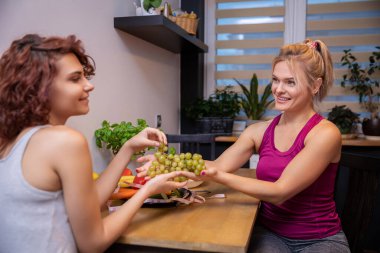 Young female friends sit at the kitchen table and hold green grapes. The woman in the foreground is blurred. On the table there are plates with vegetables and fruits. In the background is a window.