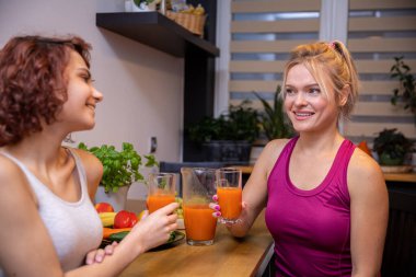 Two friends sitting in the kitchen at the table. The women in the meeting are holding glasses with orange juice. in the background a curtained window, on the table herbs in a pot.