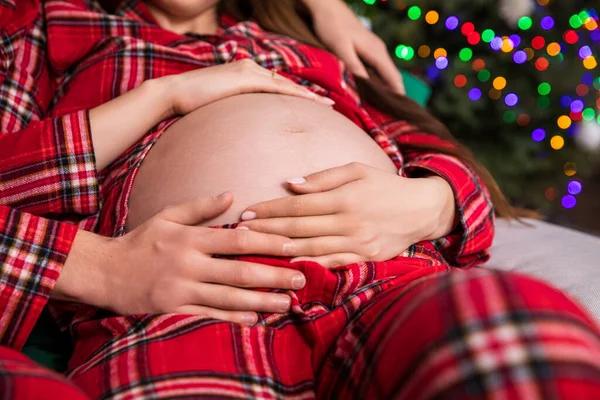 Close-up on the belly of a woman in advanced pregnancy. The man sits behind the woman and both put their hands on the pregnant belly. The parents-to-be are wearing the same red checkered pajamas.