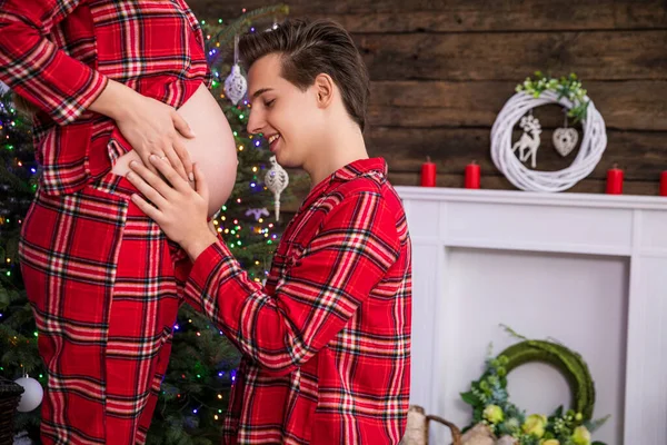 Close-up of the pregnant belly in profile. The man kneels in front of the woman looking at the pregnant belly and touching it. The couple is wearing identical red checkered pajamas.
