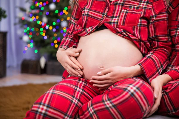 Large close-up of pregnant womans belly holding hands on her belly. A womans hands with an engagement ring are stroking her pregnant belly. Smudged Christmas lights in the background.