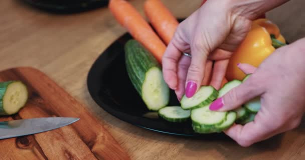 女性がキッチンで調理し 宝石や長いピンク色の爪を身に着けている間に野菜の食事を準備します スライスしたキュウリはファッショナブルな黒いプレートの上にあります — ストック動画
