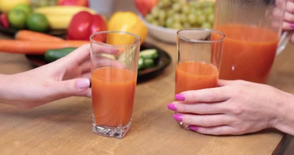 2人の女性の出会い オレンジジュースのグラスを持っているキッチンの2人の女性 背景には 茶色のテーブルの上にカラフルな野菜や果物が立っているプレート — ストック動画