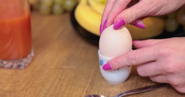 一个女人把一个煮熟的鸡蛋从它的壳里剥下 紧紧地放在她的手上 前排有粉色长钉的手 后排有一个模糊的杯子 盘子里有果汁和模糊的蔬菜 — 图库视频影像