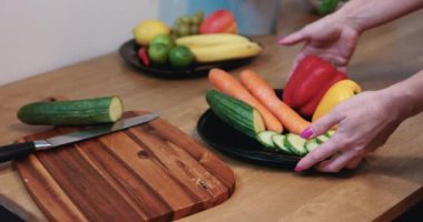 A neat womans hands present a plate full of vegetables, while a plate of fruit can be seen fuzzy in the background. Cucumber slices are neatly arranged on an elegant black plate.
