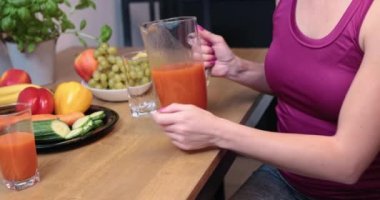 A woman sitting at a wooden table holding a glass pitcher and pouring orange juice into a glass. In the background you can see fruits and vegetables on black plates