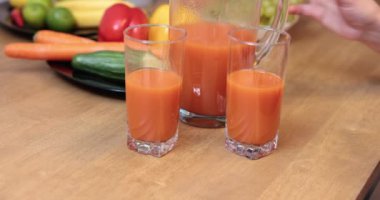 A clear glass jug filled with orange juice stands on the kitchen table. The tall glasses containing the juice are on the kitchen counter. In the background you can see fuzzy fruits and vegetables