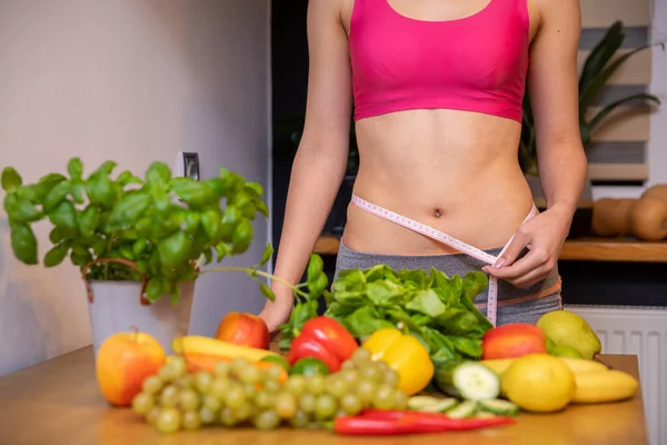 an athletic woman with a narrow waist wraps measure tape around her waist. In front of the womans figure stands a table and on it lie colorful vegetables, fruits and herbs. In the background is a
