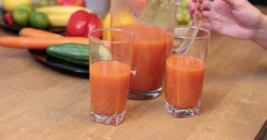A clear glass jug filled with orange juice stands on the kitchen table. The tall glasses containing the juice are on the kitchen counter. In the background you can see fuzzy fruits and vegetables