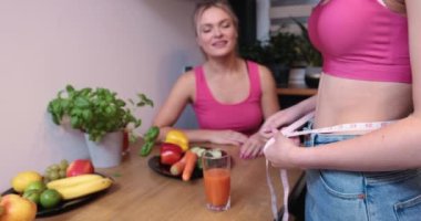 Two people in the kitchen at a table set with healthy food, fruits, vegetables. A young girl measures the circumference of her waist. A colleague admires the effects of weight loss.