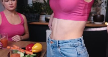 The person standing sideways shows an uncovered, slim belly against a blurry background of green potted plants standing on a wooden windowsill. The girl looks at the effects of her friends weight