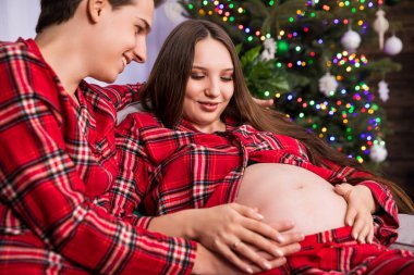 A couple expecting a baby sits on a couch near a dressed Christmas tree, which can be seen in a blurry background. The parents-to-be are wearing the same plaid pajamas.