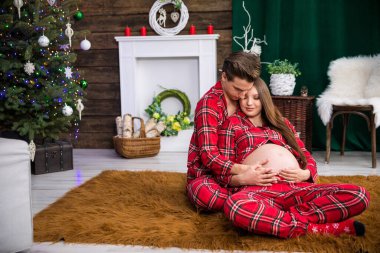 A couple of parents-to-be, hugging each other, sit on a fluffy brown carpet. The man embraces the woman from behind. The woman leans on the man.