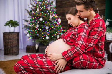 Hamile bir kadın arkasında oturan bir adama sırtını yaslar. Adam kadına sarılıyor ve elini karnının üstünde tutuyor. Kucaklaşan çift, lekeli bir Noel 'e karşı profilden görünebilir.