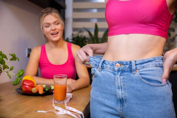 A blurry figure of a woman sitting in the background at a table looks at a friend standing in front. In the foreground, a close-up of a portion of the girls torso with her slim belly exposed.