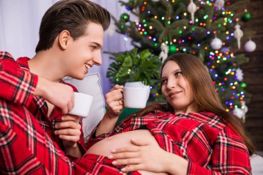 A woman in advanced pregnancy shows off her exposed pregnant belly while resting her legs on her partners knees. The couple looks into each others eyes and holds white cups in their hands.