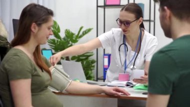 Nice and cheerful atmosphere in the doctors office during a blood pressure check. A pregnant woman and her husband sit with the doctor.