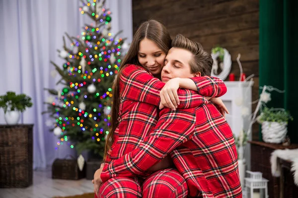 A couple dressed in identical red plaid pajamas cuddle affectionately as the woman sits on the mans lap. In the background is a smudged Christmas tree with Christmas lights.