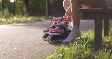 Güneşli bir parkta, beyaz çoraplı bir kadın bir çift tekerlekli paten kaldırıyor. Bir kız bacak bacak üstüne atarak parkta oturuyor. Kaldırımda güneş ışığı var..