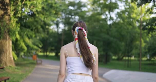 从腰部到后方看得见的人正在公园里散步 这个女孩穿着白色紧身无袖上衣 她的发型是用马尾辫扎住围巾的 她的头发很长 — 图库视频影像