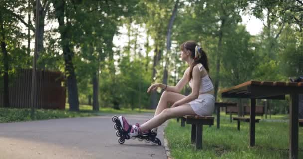 若い女性が公園のベンチに腰を下ろしている 背景には木々や芝生 緑がたくさん見られます 女の子は靴の代わりに足にローラーブレードを持っています 彼女はきついストラップレスを着ている — ストック動画
