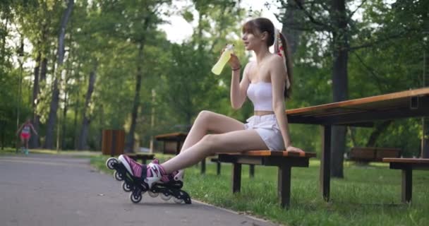 一个脚上戴着滚动式刀片的女孩放松了 一个女孩坐在公园的长椅上 她拿起一瓶黄色的饮料 开始喝酒 她笑着用手捂住脸 — 图库视频影像