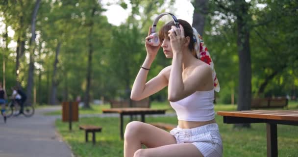 公園のベンチに座っている女の子は ワイヤレスヘッドフォンを頭の上に置きます 彼女は音楽を聴きながら微笑む 背景にはアスファルトの路地に沿って転がる人々のぼやけた姿 — ストック動画