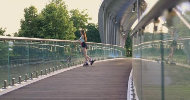 黑发姑娘站在玻璃栏杆旁边的桥上 她转过头 笑着开始滑冰 那个滑冰姑娘穿着紧身的夏季运动服 提着一个时髦包 — 图库视频影像