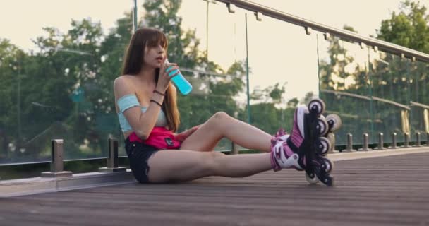 一头长发的黑发 锻炼后疲倦了 双腿直着坐着 女孩靠在桥上 背靠在玻璃透明的栏杆上 她伸手去拿一个有嘴巴的瓶子 — 图库视频影像