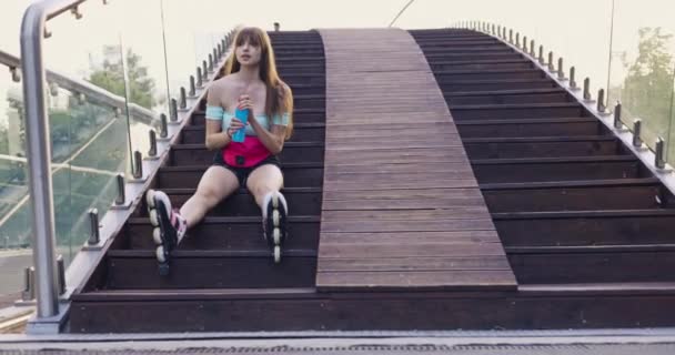 训练后 滑雪者女孩疲倦地坐在木制楼梯上 双腿挺直 她穿着旱冰鞋和紧身运动服 她伸手去拿一个瓶子 喝了一杯蓝色的伏特加 — 图库视频影像