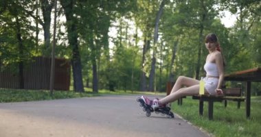 Ayakları patenli bir kız parkta bir bankta oturuyor ve rahatlıyor. Kız bir şişe sarı izotonik içecek aldı. Kadın gülümsüyor..