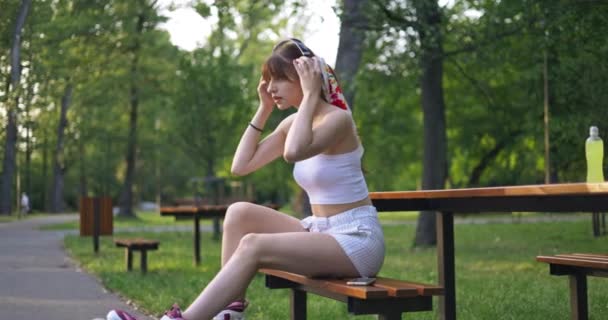 公園のベンチに座っている若い人 彼女はワイヤレスヘッドフォンを頭の上に置きます 女の子の隣には黄色い等方性ドリンクのボトルが立っています 背景には女性のぼやけた姿 — ストック動画