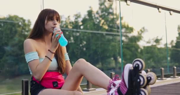一个人在锻炼后喝了一杯冰醋酸饮料 女孩闭上了瓶子的嘴 一个女孩坐在桥上 训练后休息 微笑着向前看 在她的脸上进行了特写 — 图库视频影像