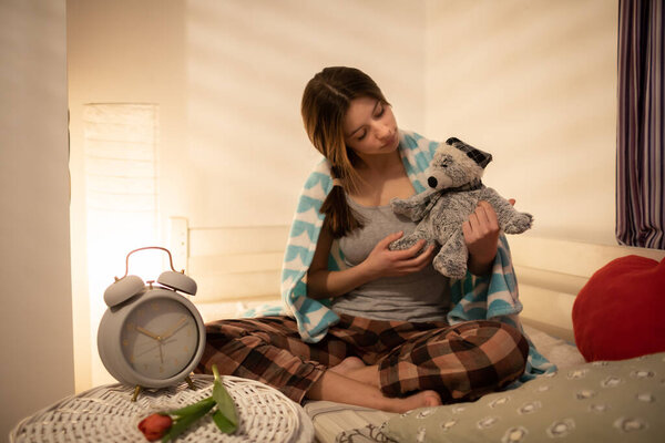 The girl holds the cuddly teddy bear in her hands. Bright bedroom with decorations. The light of a lamp on the wall.