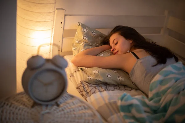 A woman with a slight smile sleeps on her stomach in her bed. A large alarm clock stands on the bedside table.