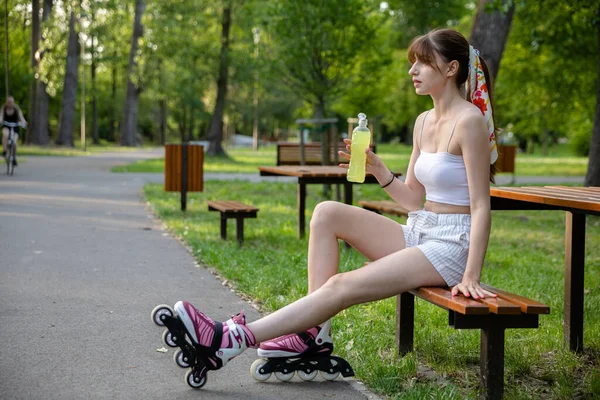 短パンの少女とスポーツのトップがベンチに座っている 彼女は黄色い等方性ドリンクのボトルを持っている 背景には自転車に乗る姿が見える ぼやけた木々や芝生 — ストック写真