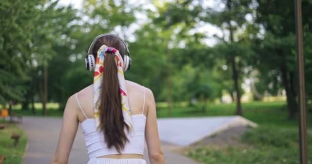 在一个沿着公园小巷移动的女孩的后面特写 在炎热的夏日散步的女孩 这个女孩穿着一件白色运动服 无袖上衣走路 女孩穿着一条缠着围巾的马尾辫 — 图库视频影像