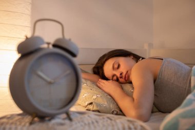 The girl sleeps with her head on a large pillow. The light of a lamp on the wall. clipart