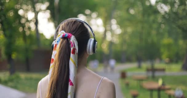 穿着马尾辫的女孩的后背特写 这个女孩正在公园里散步 头上戴着大大的无线耳机 许多人从街对面经过她 — 图库视频影像
