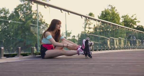 緩やかな髪の長い髪のブルネットが橋の上に座っています 彼女はガラスの透明な手すりに背を向けている 靴の代わりに彼女の足にはローラーブレードがあります 彼女は横になって — ストック動画