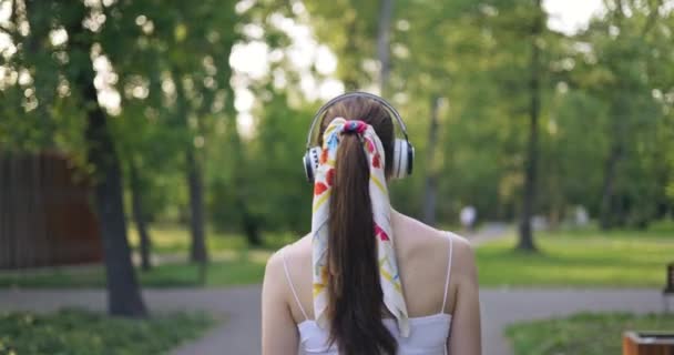 一个穿着马尾辫 裹着围巾的女孩的后背特写 这个女孩正在公园里散步 头上戴着大大的无线耳机 许多人从街对面经过她 — 图库视频影像
