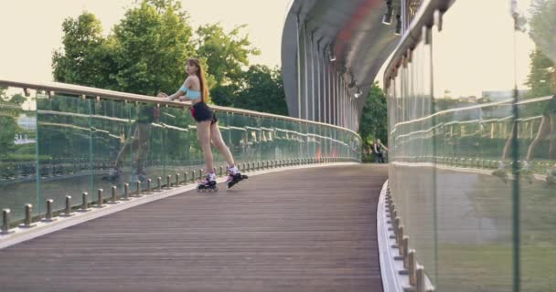 一个滑冰姑娘在桥上的木制表面上滑冰 桥与玻璃栏杆 女孩在笑在这个背景下 可以看到别人走路和骑自行车的模糊形象 — 图库视频影像