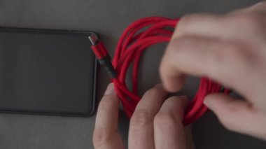 Gri bir bezin üzerinde duran cep telefonunun yakın çekimi. Telefonun yanında uzun kırmızı kablosu olan bir şarj aleti var. İnsan eli kabloyu çözer ve mikro fişi telefon prizine takar.