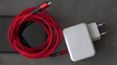 Bir insan eli kırmızı bir kabloya bağlı beyaz bir şarj aletine uzanır. Şarj aleti ve kablo cep telefonunun üstünde. İnsan eli şarj aletini ve kabloyu alır ve alır. El o zaman.