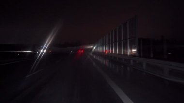 Araba asfalt bir yol boyunca ses engellerini aşarak ilerliyor. Sokak lambalarından ve trafik ışıklarından gelen bulanık ışıklar. Şehrin bir sokağında cam silecekleri açık bir araba kullanmak. Sokak manzarası.