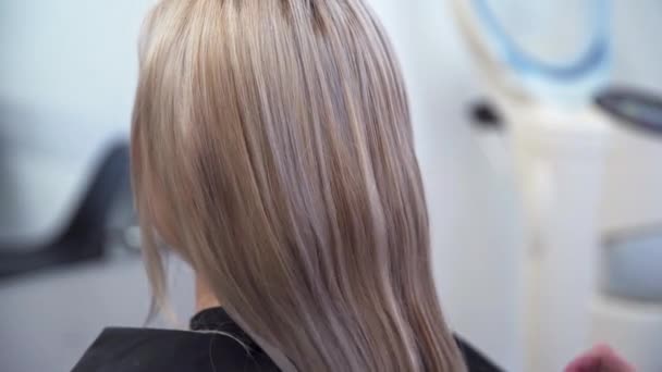 美容師は コンディショナーでスプレーすることで さらなる治療のためにクライアントの髪を準備します 後ろから見えるブロンドの髪の女性の頭 前景では 美容師の手 — ストック動画