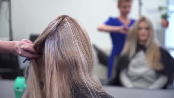 美容師は 顧客の髪をストランドに分離し ヘアピンでそれらをピン 床屋の椅子に座っている女性 鏡像で見た女性と美容師 美容師は青い服を着ている — ストック動画