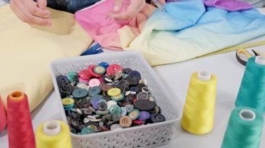 Önce karıştırıyor ve sonra iki eline de farklı renkte düğmeler alıyor. Terzi atölyesini küçük aksesuarlarla destekliyor..